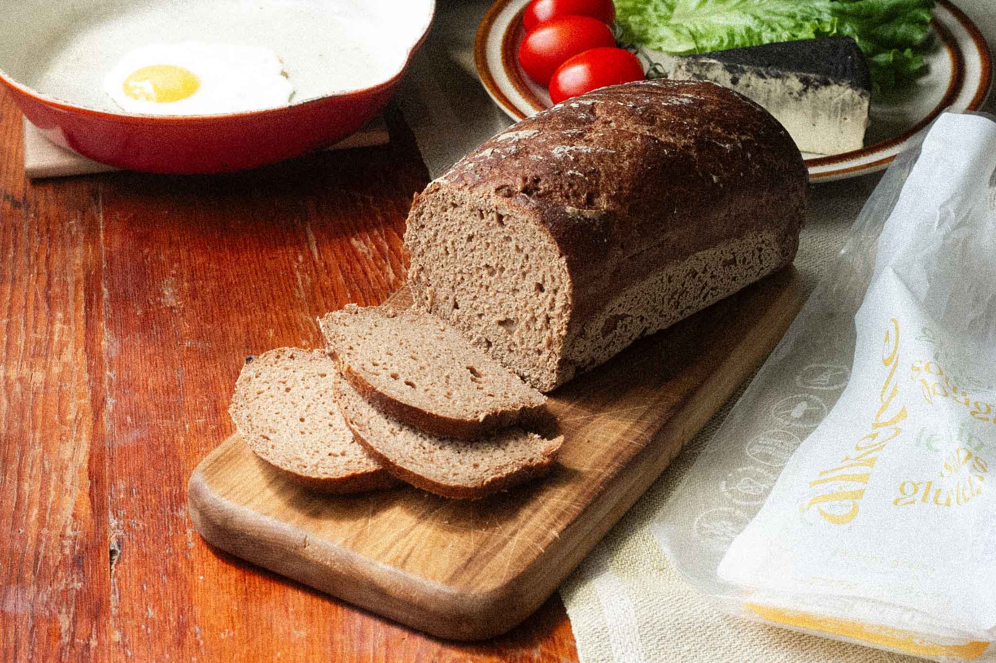 alkeme's scrumptious ancient grains loaf sitting on a cutting board with a few slices already cut exposing the delicious sourdough crumb and air pockets within. Behind the laof cashew cheese, tomatoes, lettuces and an egg in a frying pan are waiting to be topped on an empty slice. To the right is the yellow, white, and green packaging of the ancient grains loaf. 