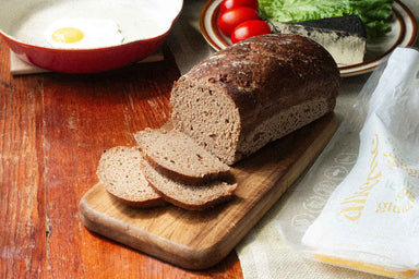 alkeme's scrumptious ancient grains loaf sitting on a cutting board with a few slices already cut exposing the delicious sourdough crumb and air pockets within. Behind the laof cashew cheese, tomatoes, lettuces and an egg in a frying pan are waiting to be topped on an empty slice. To the right is the yellow, white, and green packaging of the ancient grains loaf.