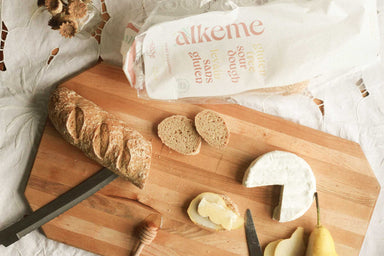 An image of an alkeme demi baguette on a wooden cutting board with a few plain slices and one slice covered in pear, cheese, and honey. Alkeme packaging in the top part of the frame.
