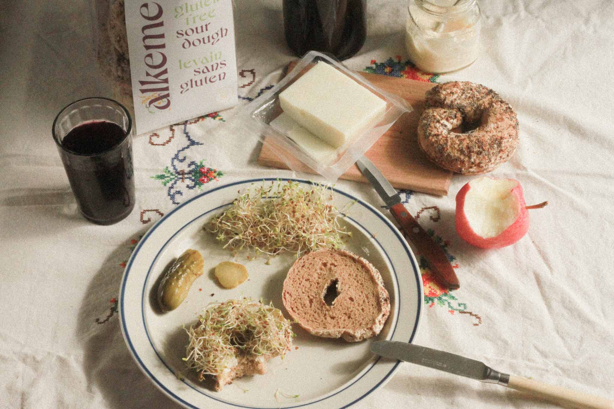 Image of alkeme Everything Bagel, one side covered with cheese and sprouts and the other waiting to be topped. A pickle on the plate and cheese, mayo and grape juice in the background. A half eaten apple off to the side and bagel packaging in the background.