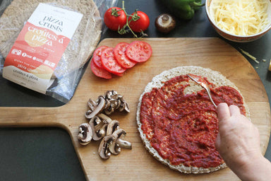An image of alkeme's delicious pizza crust being lathered with hommade herby tomato sauce on a classic wooden pizza board. Sliced mushrooms, sliced tomatoes and grated cheese and a few scattered veggies frame the scene.