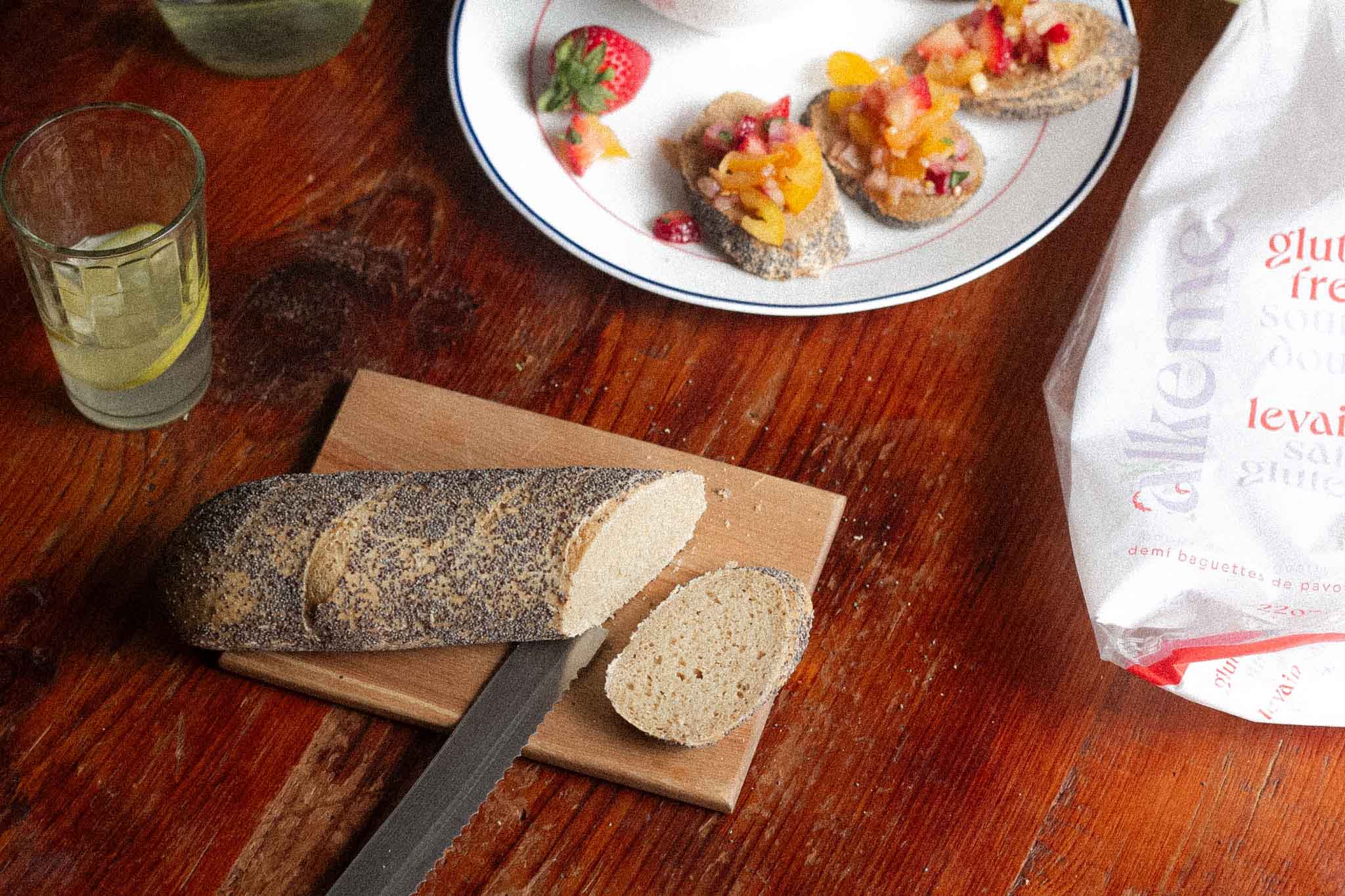 An image of an alkeme Poppy Seed Demi Baguette being sliced on a cutting board and a plate with strawberry bruscettan topped slices in the background. A glass of lemon water off to the left and the alkeme Poppy Demi Baguette packaging off to the right.