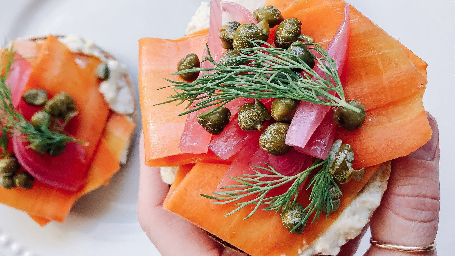 Hand holding Faux Lox Bagel with birghtly coloured smoked carrot, capers, pickled red onion, and fresh dill