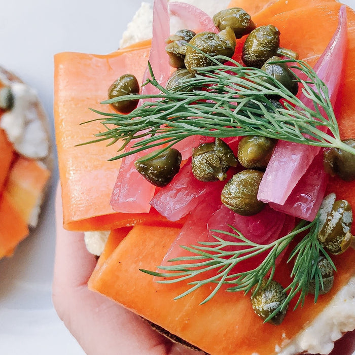 Hand holding Faux Lox Bagel with birghtly coloured smoked carrot, capers, pickled red onion, and fresh dill