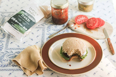 An image of alkeme's Burger Bun sandwhiching a plant based burger with sliced tomatoes, homemade ketchup and relish in mason jars in the background. Alkeme Burger Bun packaging in the top left of the frame.