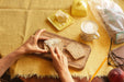 Image of hands spreading butter on two pieces of alkeme country Loaf Bread on a yellow table cloth with packaging in the top right corner of the frame and a delicious foamy coffee waiting to be drunk.