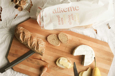 An image of an alkeme demi baguette on a wooden cutting board with a few plain slices and one slice covered in pear, cheese, and honey. Alkeme packaging in the top part of the frame. 
