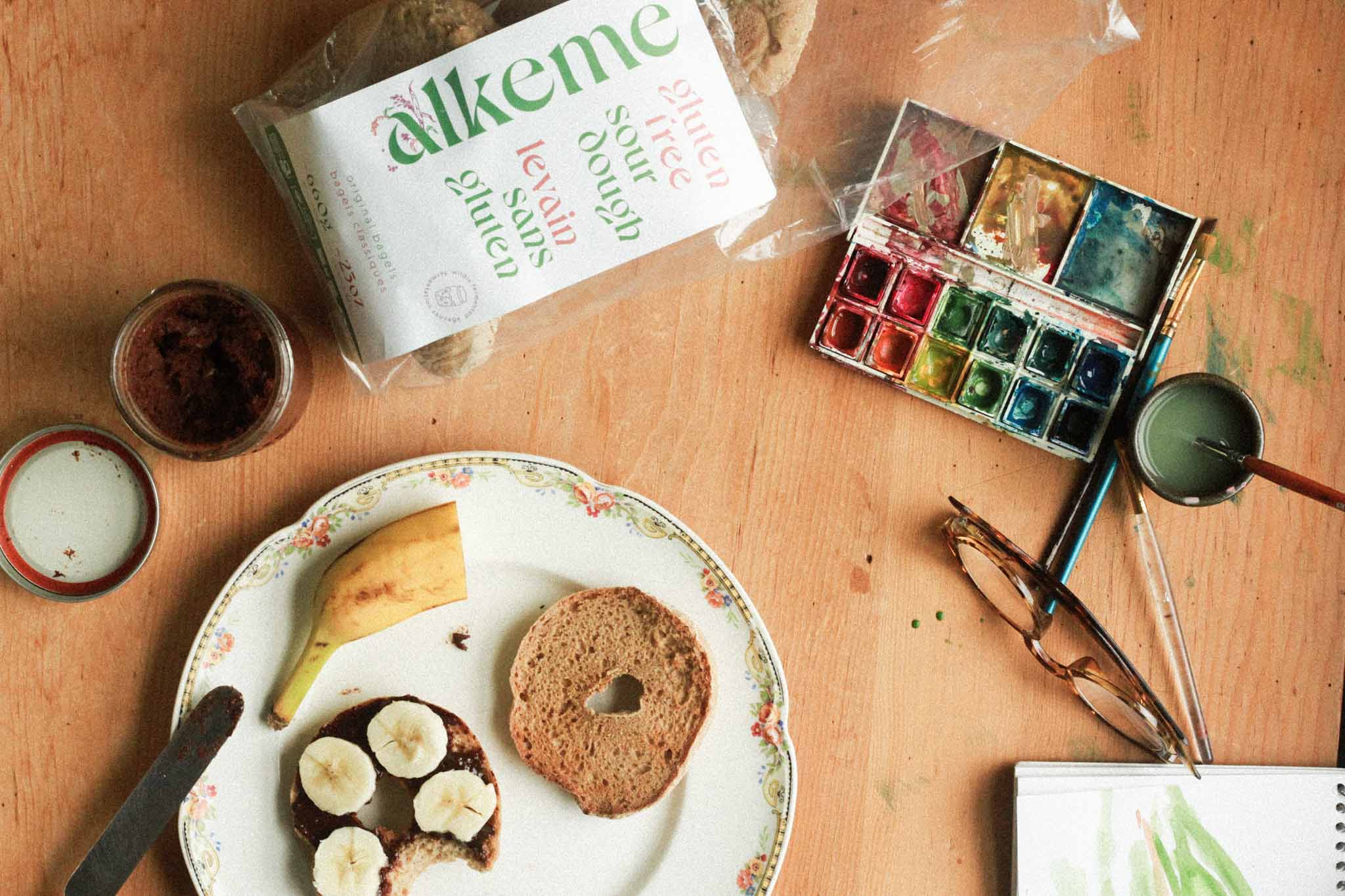 An image of alkeme Original Bagel, one half topped with homemade nutella and banana slices and the other half waiting to be topped. Paints and a sketch book and glasses off in one corner and alkeme bagel packaging at the top of the frame.