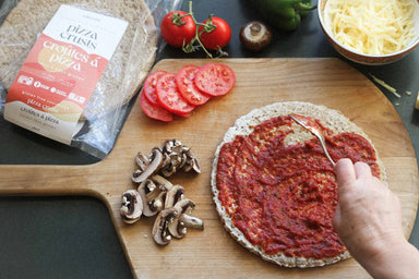 An image of alkeme's delicious pizza crust being lathered with hommade herby tomato sauce on a classic wooden pizza board. Sliced mushrooms, sliced tomatoes and grated cheese and a few scattered veggies frame the scene. 