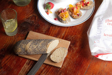 An image of an alkeme Poppy Seed Demi Baguette being sliced on a cutting board and a plate with strawberry bruscettan topped slices in the background. A glass of lemon water off to the left and the alkeme Poppy Demi Baguette packaging off to the right.